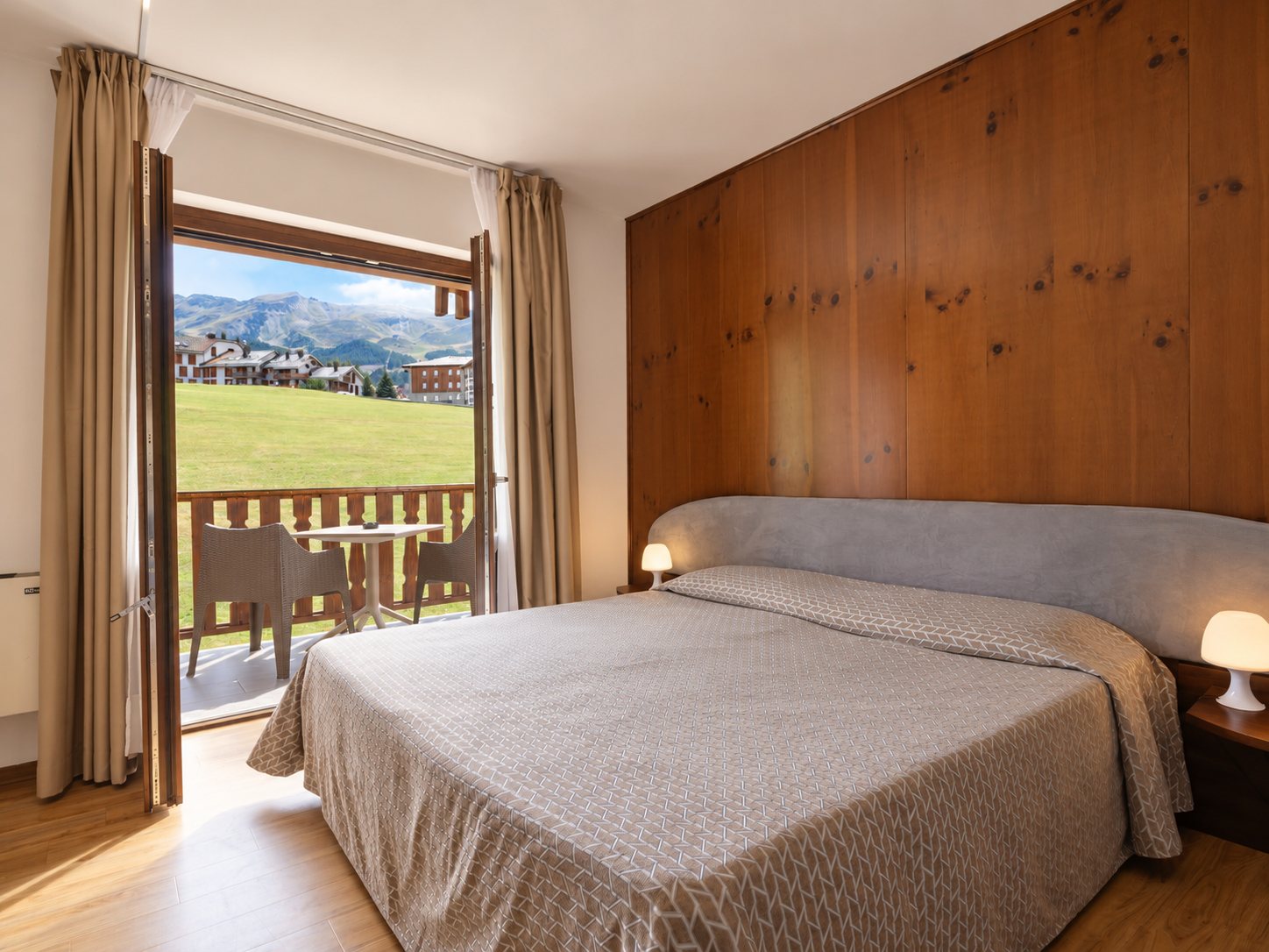 Camera con letto matrimoniale, parete in legno e portafinestra aperta sul balcone con vista sui prati di Pila