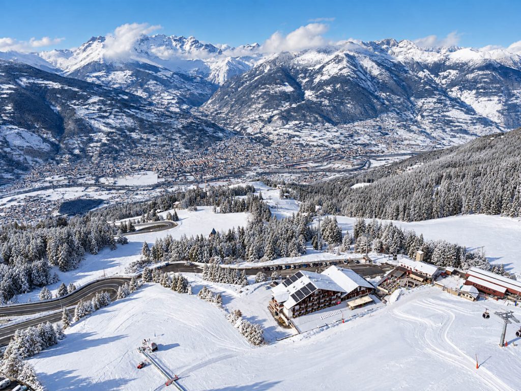 Vista dall'alto dell'hotel a Pila d'inverno, tra le piste e i boschi innevati, con la valle di Aosta sullo sfondo