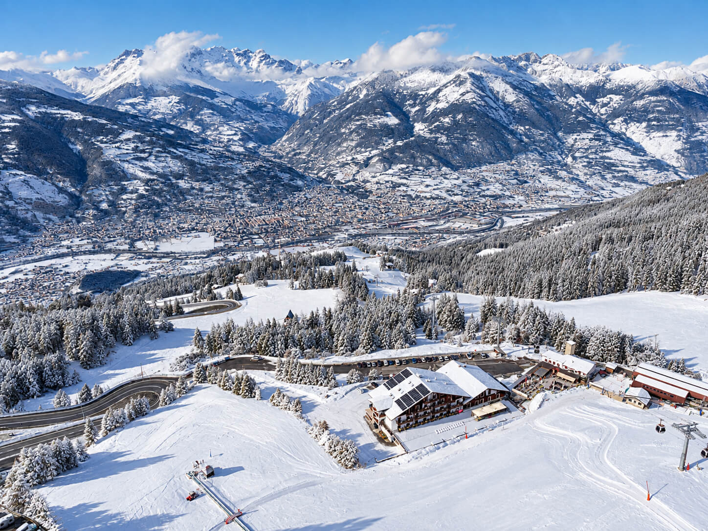 Vista dall'alto dell'hotel a Pila d'inverno, tra le piste e i boschi innevati, con la valle di Aosta sullo sfondo